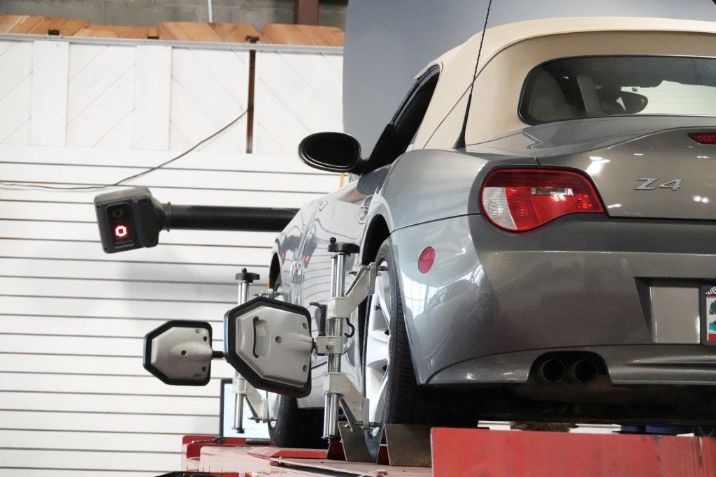 Silver BMW Z4 convertible on an alignment rack with sensors clamped to the wheels while a computerized alignment camera points at the rear of the car in a repair shop.