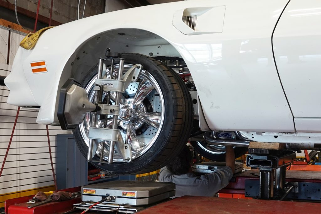 White muscle car on a lift with its front wheel clamped in a computerized alignment machine while a mechanic works underneath on upgraded suspension and brakes.